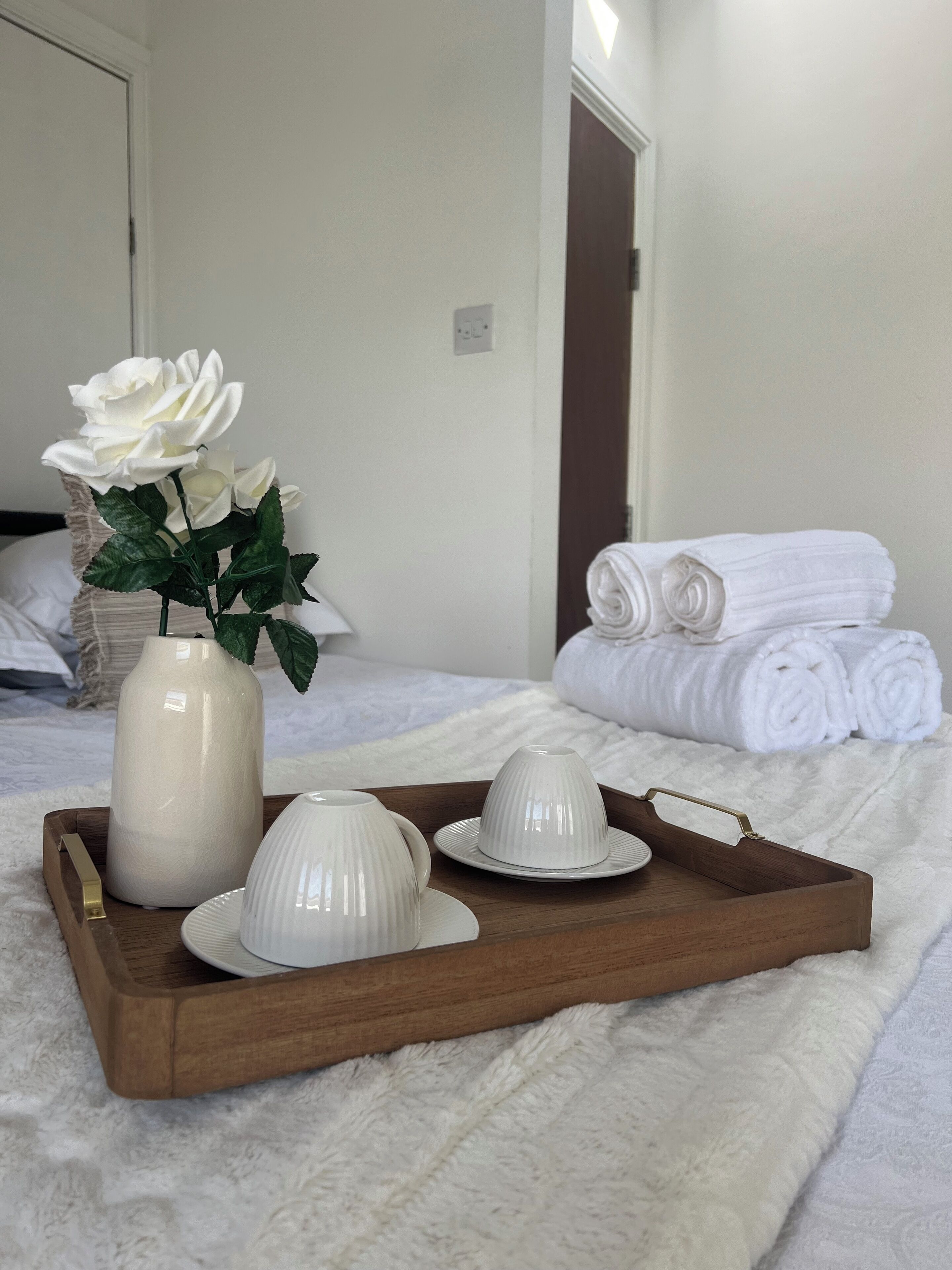 A hotel room with a neatly made bed, a tray with white teacups and a vase of white roses, and rolled white towels.