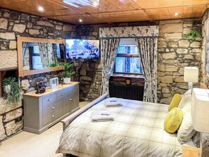Ein Schlafzimmer mit Steinwand, Holzboden, einem Bett mit Karomusterdecke, einem Nachttisch mit Lampe und einem Fernseher an der Wand.