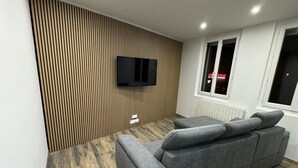 A modern living room with a grey sofa, wooden flooring, a mounted TV, and a window with a view. - Appartement Cosy (Roussillon)