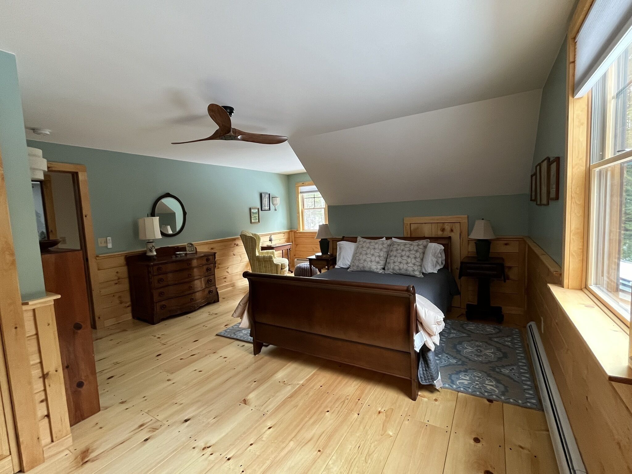 A bedroom with a wooden floor, a bed with a headboard, a dresser, a chair, a ceiling fan, and a window.