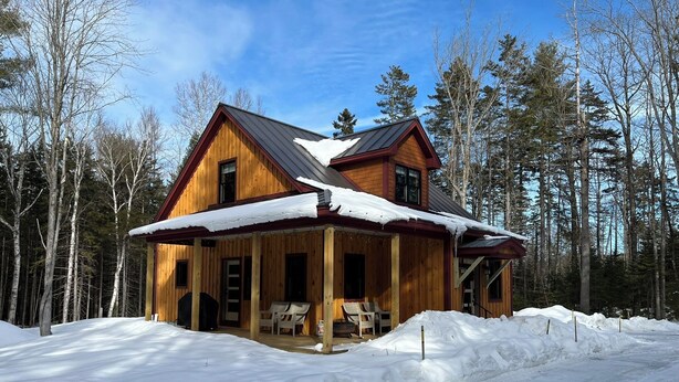Una cabaña de madera con un porche, rodeada de nieve y árboles.