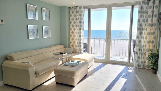 A living room with a beige sofa, a small table, and a potted plant. There are framed pictures on the wall and a view of the sea through the balcony doors. - Looking for More than Two Bedrooms on the Beach? (Marco Island)