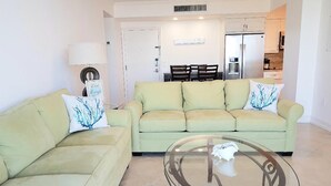 A living room with two light green sofas, a glass coffee table, and a dining area in the background. - Looking for More than Two Bedrooms on the Beach? (Marco Island)