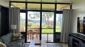 Ein Wohnzimmer mit Sofa, Couchtisch und Fernseher, große Fenster mit Blick auf einen Garten und Palmen.