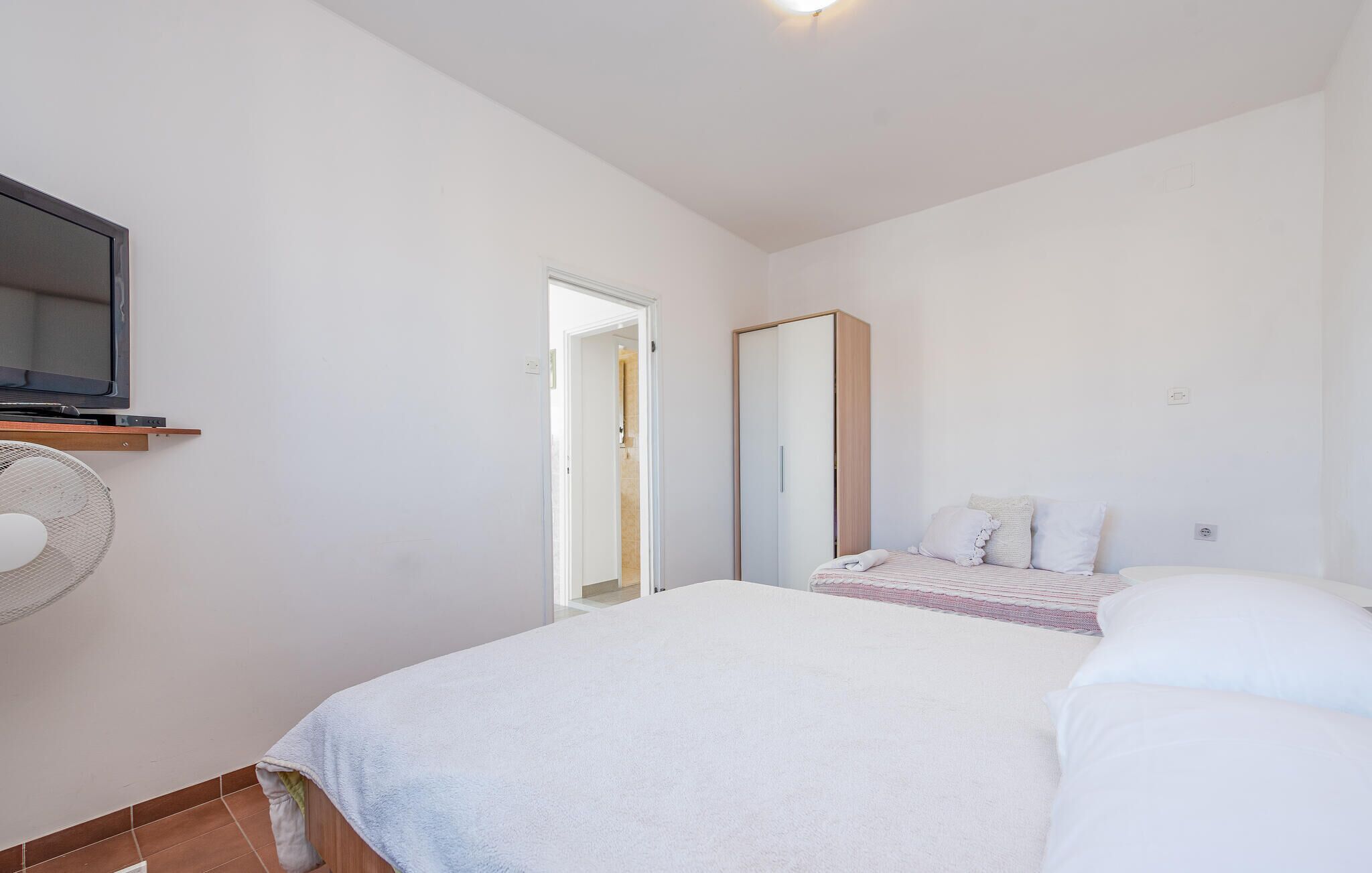 Quarto com cama, televisão presa a uma prateleira, guarda-roupa e porta que dá para outro espaço.