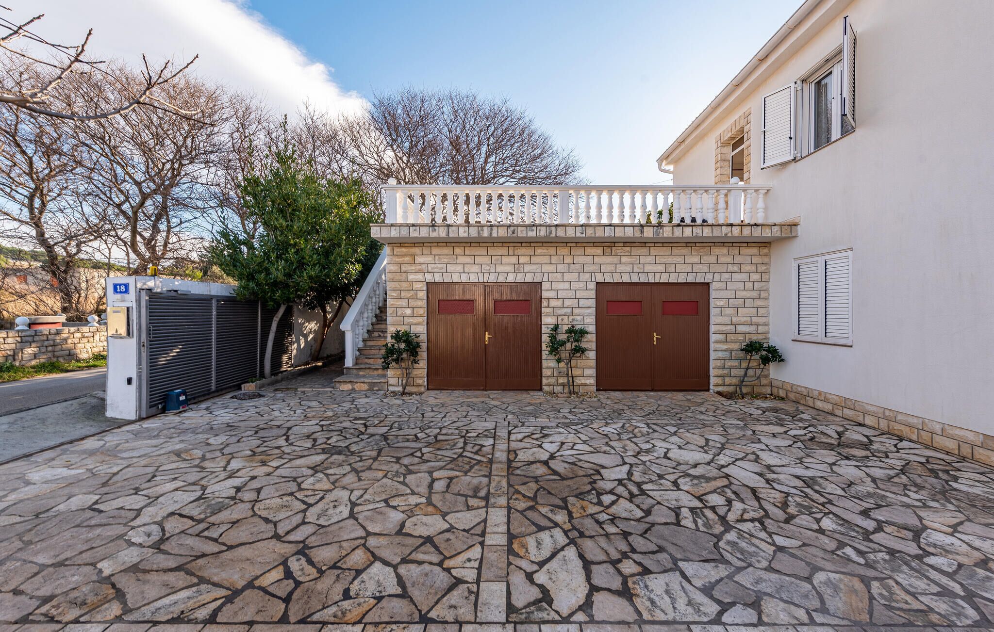 Un patio empedrado con dos puertas de madera, un acceso con portón y una pequeña escalera que lleva a un balcón.