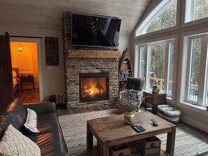 A cozy living room with a stone fireplace, a flat-screen TV, a comfortable sofa, and a large window offering a view of the outdoors.