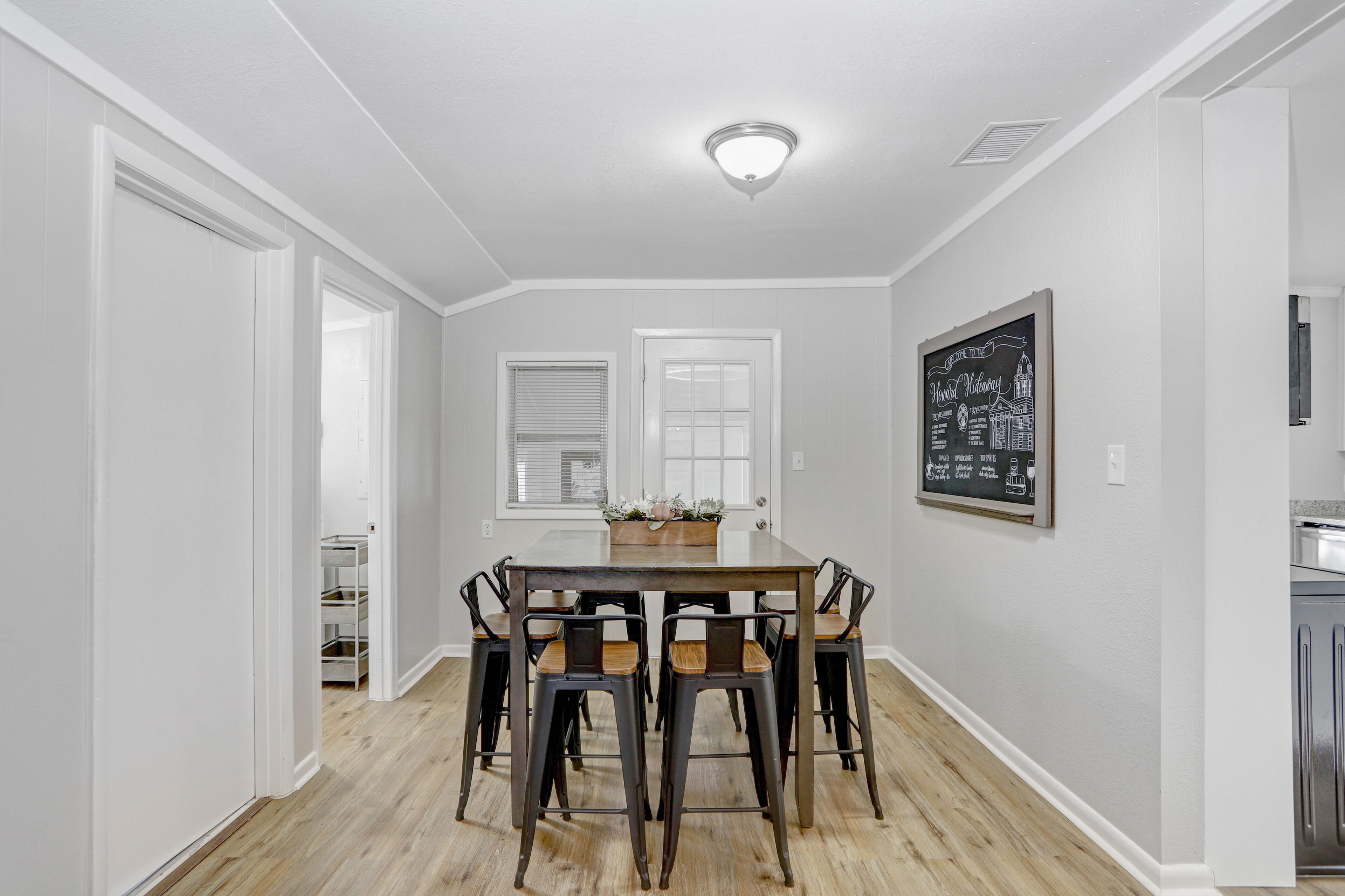 Un coin repas avec une table rectangulaire entourée de six chaises, un plafond blanc avec un éclairage encastré et un tableau noir encadré accroché au mur.