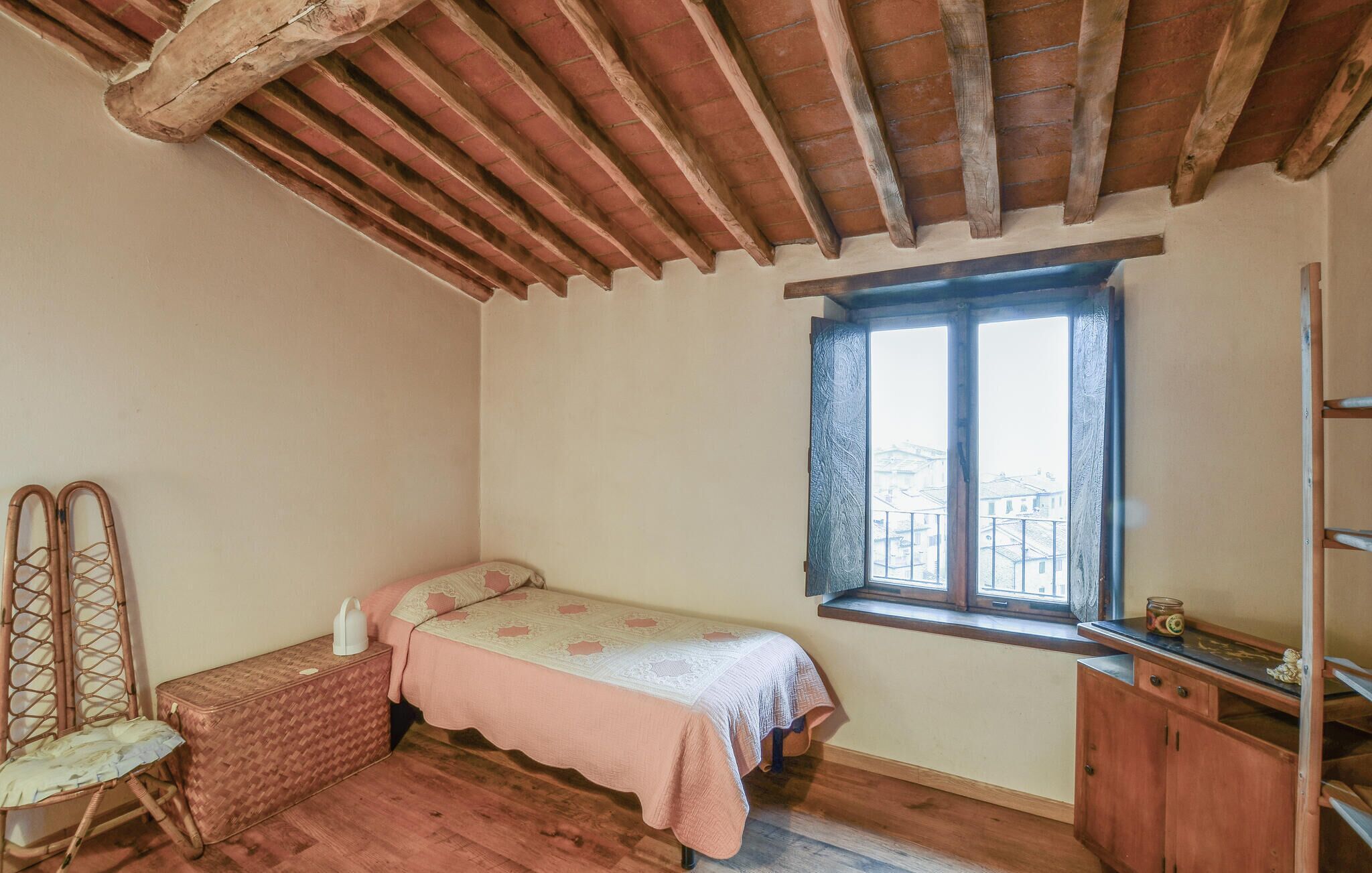 Ein kleines, gemütliches Zimmer mit einem Einzelbett, einem Holzstuhl, einem kleinen Tisch und einem Fenster mit Blick auf Gebäude.