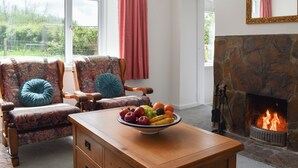 Ein Wohnzimmer mit Kamin aus Stein, zwei Sesseln mit floralem Bezug, einem Holztisch mit Obstschale und einem großen Fenster mit rosafarbenen Vorhängen.