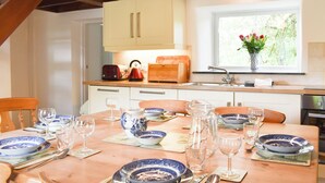 Ein Essbereich mit Holztisch, gedeckt für eine Mahlzeit, eine Küche mit Holzschränken, ein Fenster mit Blumen und ein Waschbecken.