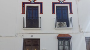 A two-story building with a white facade, two balconies with black railings, and a brown double door. - Casa Jaro en el Centro del Pueblo (Sierra de Yeguas)