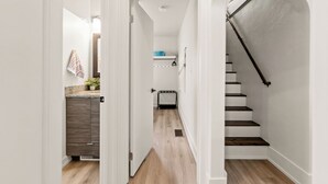 A narrow hallway with a wooden floor, white walls, and a staircase with a black handrail. - M2 -Relaxed Beach Stay • Fenced Yard, Fire Pit & 2 Living Areas, Pets welcome (Muskegon)