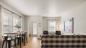 A modern living room with a checkered sofa, a dining area with a wooden table and metal chairs, and a kitchen visible in the background. - M2 -Relaxed Beach Stay • Fenced Yard, Fire Pit & 2 Living Areas, Pets welcome (Muskegon)