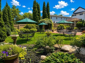 Un patio exterior bien cuidado con varias mesas y sillas, rodeado de abundante follaje y plantas con flores.