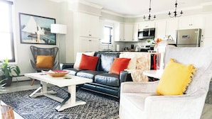 Een moderne woonkamer met een zwarte leren bank, een witte fauteuil en een salontafel. Achtergrond is een keuken met witte kasten en roestvrijstalen apparatuur.