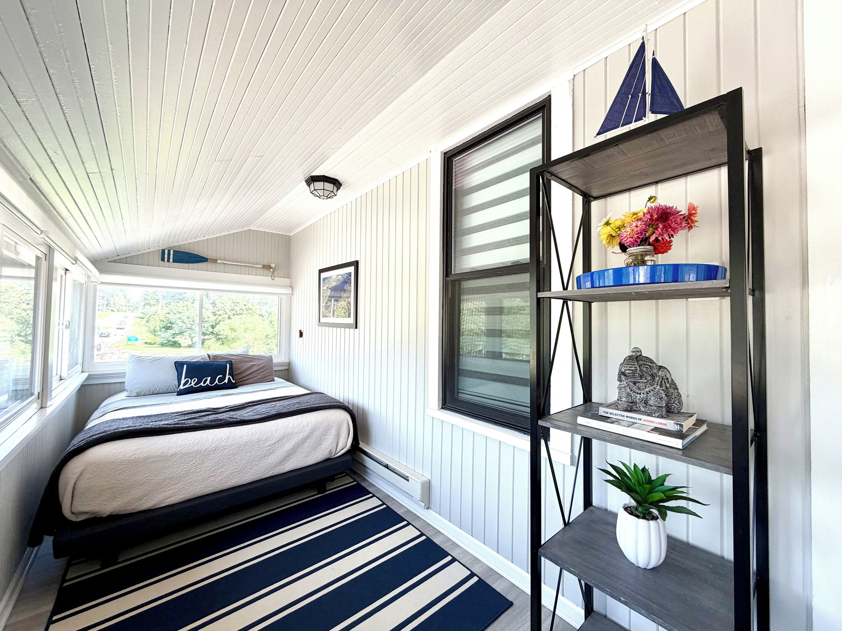 A bedroom with a bed, a window, a shelf with decorative items, and a potted plant.