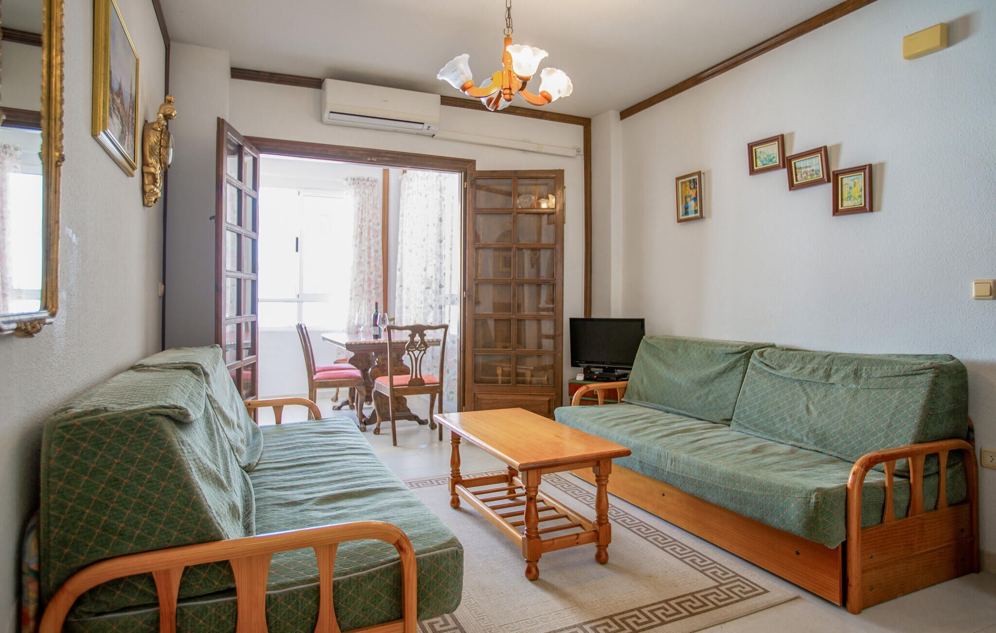 A living room with a sofa, a wooden coffee table, and a TV. There is a dining table with chairs and a window with curtains.