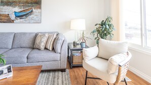 Ein Wohnzimmer mit grauem Sofa, einem Holztisch, einem weißen Sessel, einer Lampe, einer Pflanze und einem Bild eines Bootes an der Wand.