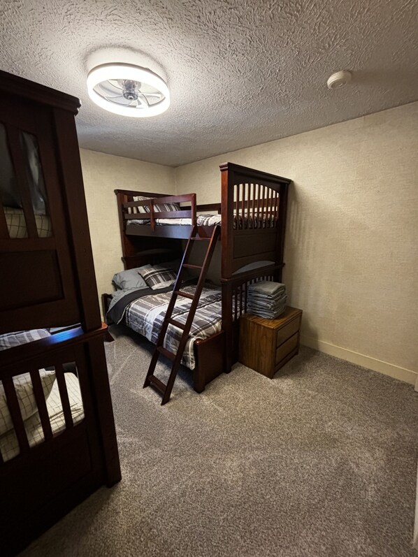 Un dormitorio con literas, una escalera de madera, un pequeño cofre de madera y un ventilador de techo.