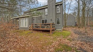 A modern cabin with a large deck and a wooden fence, surrounded by trees and a leaf-covered ground.