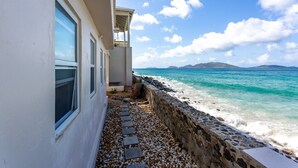 A coastal building with a stone pathway leading to the sea, a balcony, and a view of the ocean and distant mountains. - Ocean Mist - AppleSurf! Beachfront one bedroom in Apple Bay (West End Tortola)