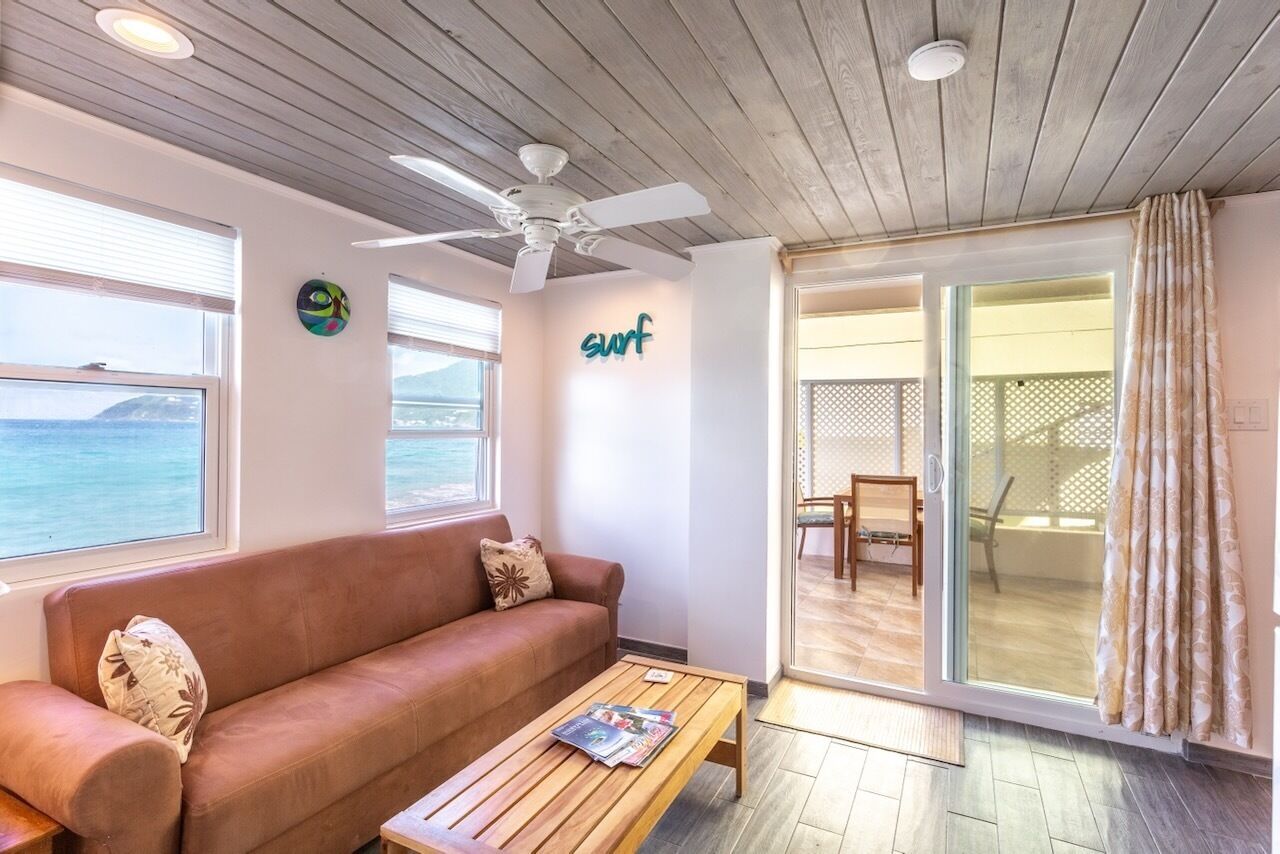 Una habitación con techo de madera, ventilador de techo, sofá, mesa de centro y vista al mar a través de amplias ventanas.