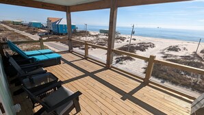 Una terraza de madera con sillones de descanso y una mesa de vidrio, con vistas a una playa que tiene casas y un muelle.
