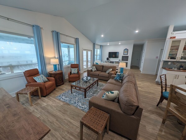 A spacious living room with a large sofa, armchairs, a coffee table, and a TV. There are large windows with curtains, a kitchen area, and a dining table.