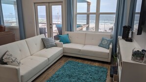 Un salón con un sofá modular blanco, un tapete azul verdoso y ventanas amplias que dan a la playa.