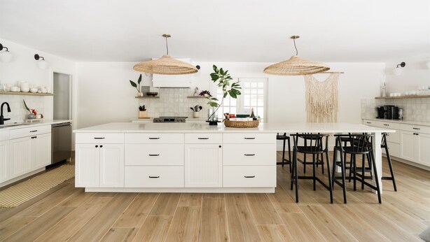 Una cocina moderna con armarios blancos, una isla central con taburetes altos y dos lámparas de mimbre colgantes.