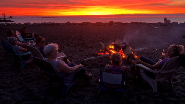Un gruppo di persone sedute attorno a un fuoco da campo su una spiaggia al tramonto.