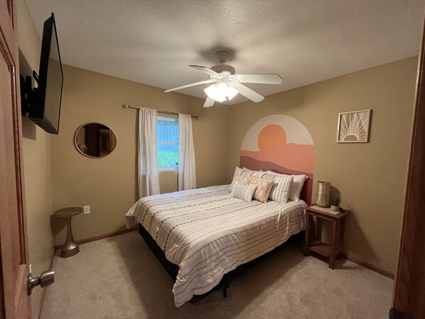 A bedroom with a bed, a ceiling fan, a window with curtains, a wall-mounted mirror, and a decorative wall piece.