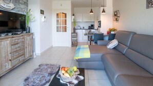 A modern living room with a grey sofa, a flat-screen TV, a wooden cabinet, and a dining area with a table and chairs. - Green Garden XVXI 205 with pool view by Interhome (De Haan)