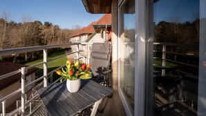 A balcony with a potted plant, a table, and a view of a building with a red roof. - Green Garden XVXI 205 with pool view by Interhome (De Haan)