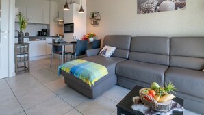 A modern living room with a grey sofa, a small coffee table with fruit, and a dining area with a kitchen in the background. - Green Garden XVXI 205 with pool view by Interhome (De Haan)