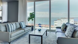Un salón moderno con sofá, una mesa de centro y un amplio ventanal con vistas al mar y edificios.