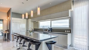 Una cocina moderna con una isla grande, taburetes de bar, iluminación colgante y vistas del cielo a través de amplios ventanales.
