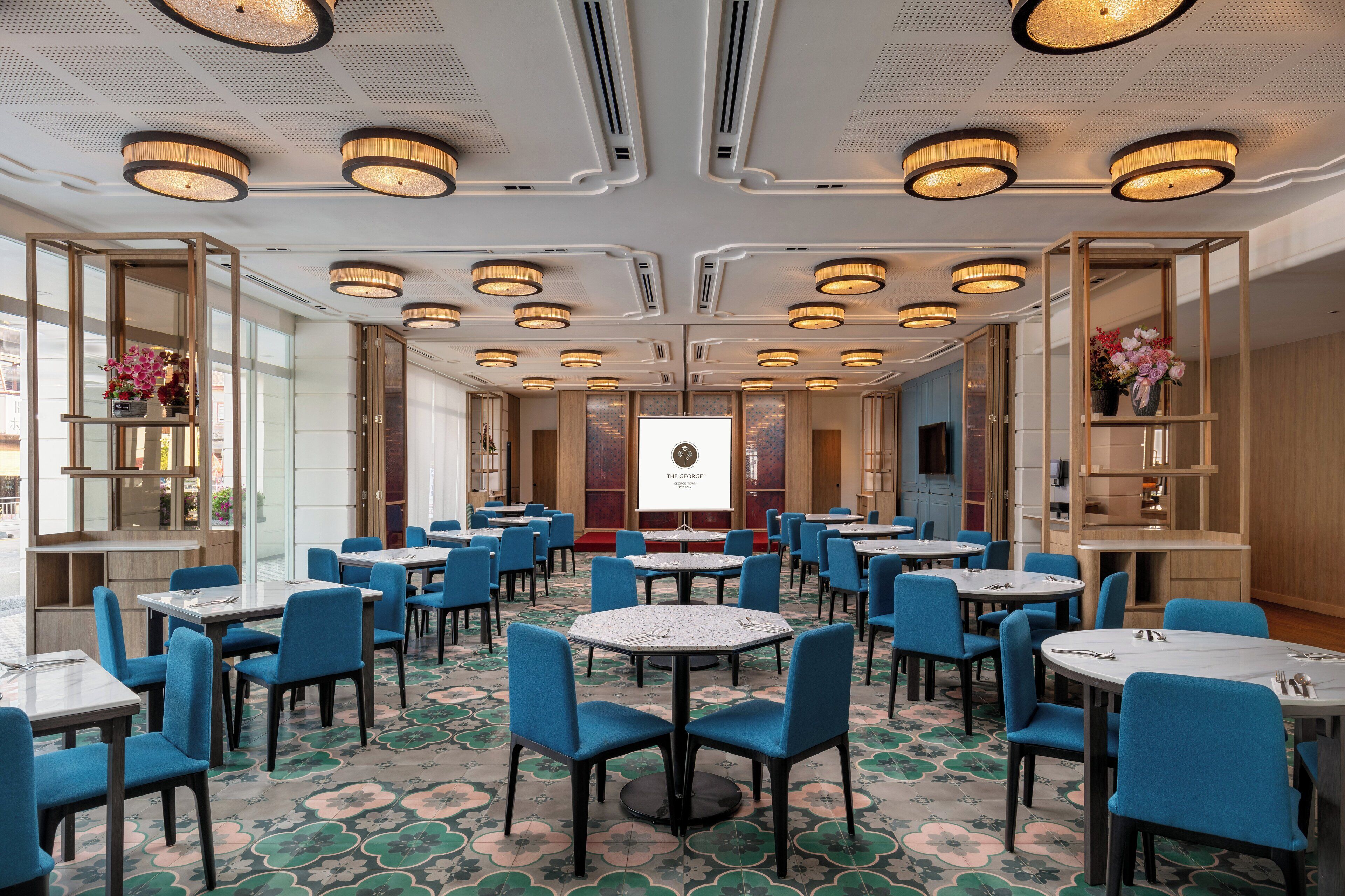 A hotel conference room with multiple tables and chairs, a large screen, and decorative shelves with flowers.