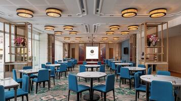 A hotel conference room with multiple tables and chairs, a large screen, and decorative shelves with flowers.