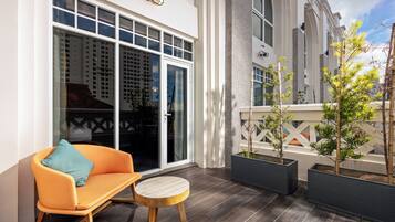A balcony with a rocking chair and a small round table, a white building with large windows, and potted plants.