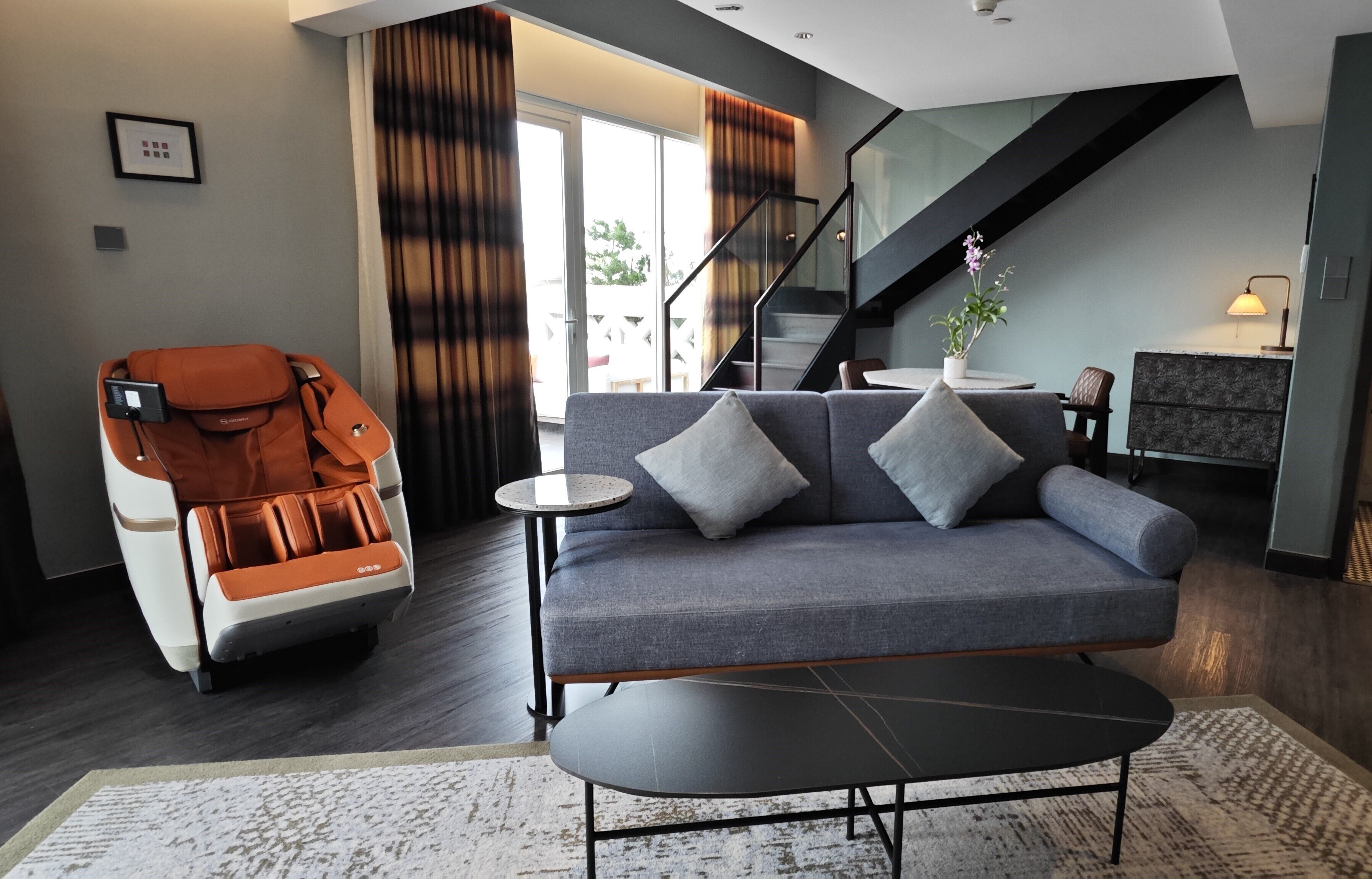 A modern living room with a gray sofa, a massage chair, a small round table, and a staircase leading to an upper level.