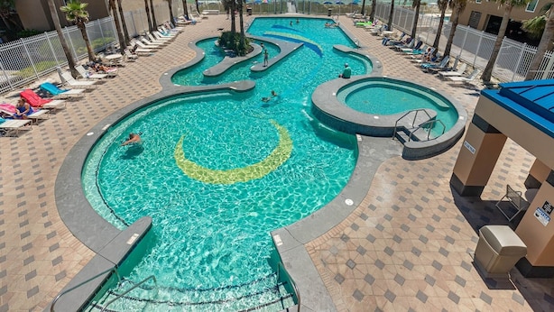 A multi-level swimming pool with a crescent moon design, surrounded by palm trees and lounge chairs. - Crystal Tower 1102 2 Bedroom Condo (Gulf Shores)