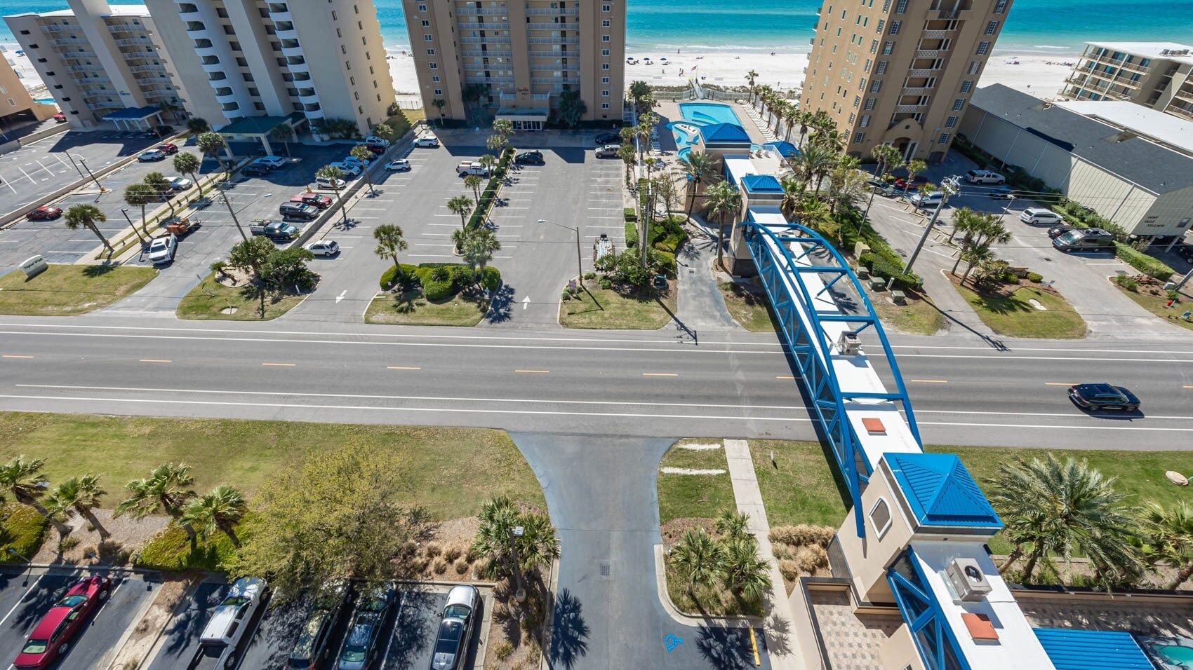 An aerial view of a coastal hotel area with a parking lot, a pedestrian bridge, and beachfront views.