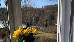 Une fenêtre avec un vase de fleurs jaunes, une table en bois sur laquelle se trouve une assiette de nourriture, et deux tasses.