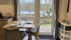 Un coin repas avec une table dressée pour deux personnes, une chaise, un buffet et une vue sur des arbres et un bâtiment à l’extérieur.