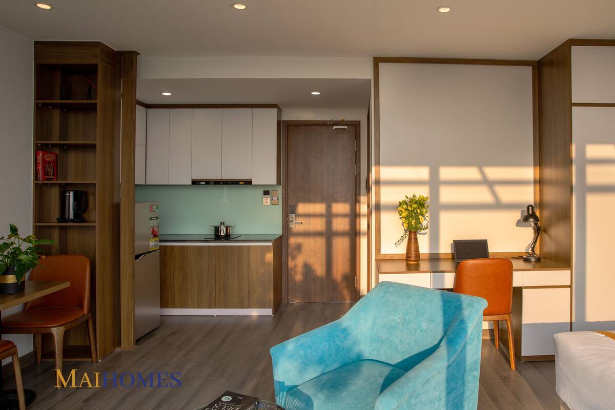 A modern hotel room with a kitchenette, a dining area with a blue sofa, and a workspace with a desk and chair.