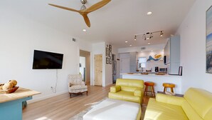 A modern living room with a yellow sofa, a white ottoman, a wooden ceiling fan, and a kitchen in the background. - Walk to Sandfest , Shared Pool, Board Walk to Beach (Port Aransas)