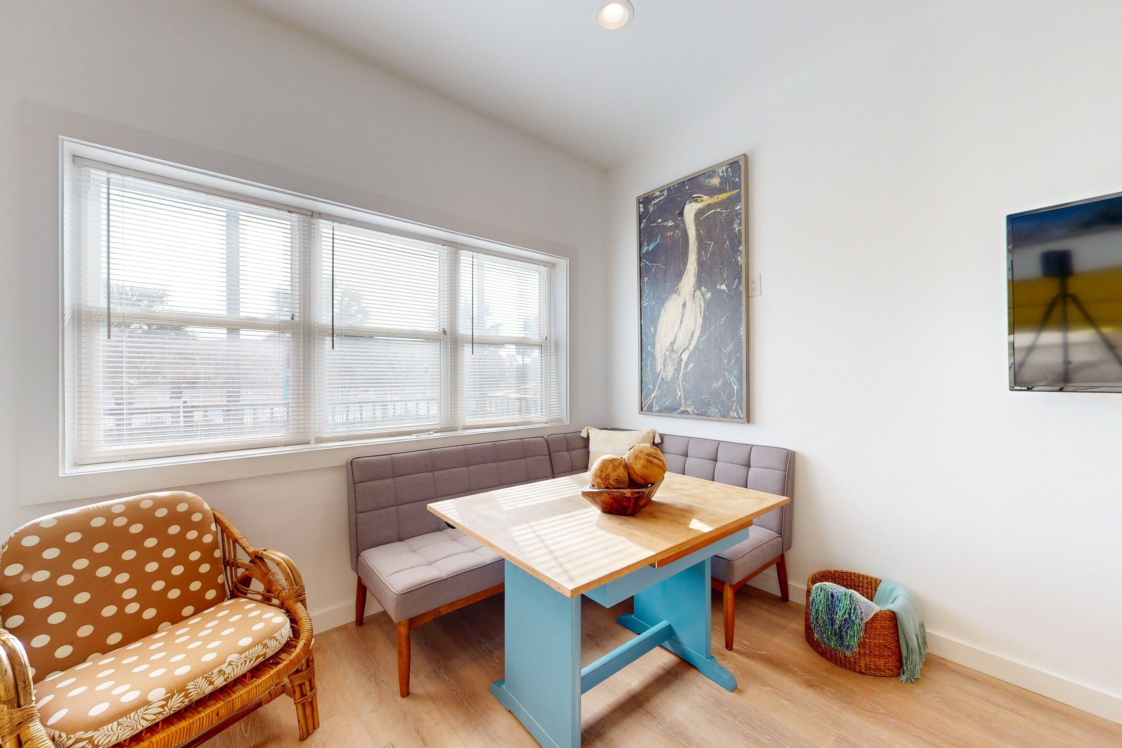 A room with a wooden table, a grey sofa, a wicker chair, and a window with blinds.