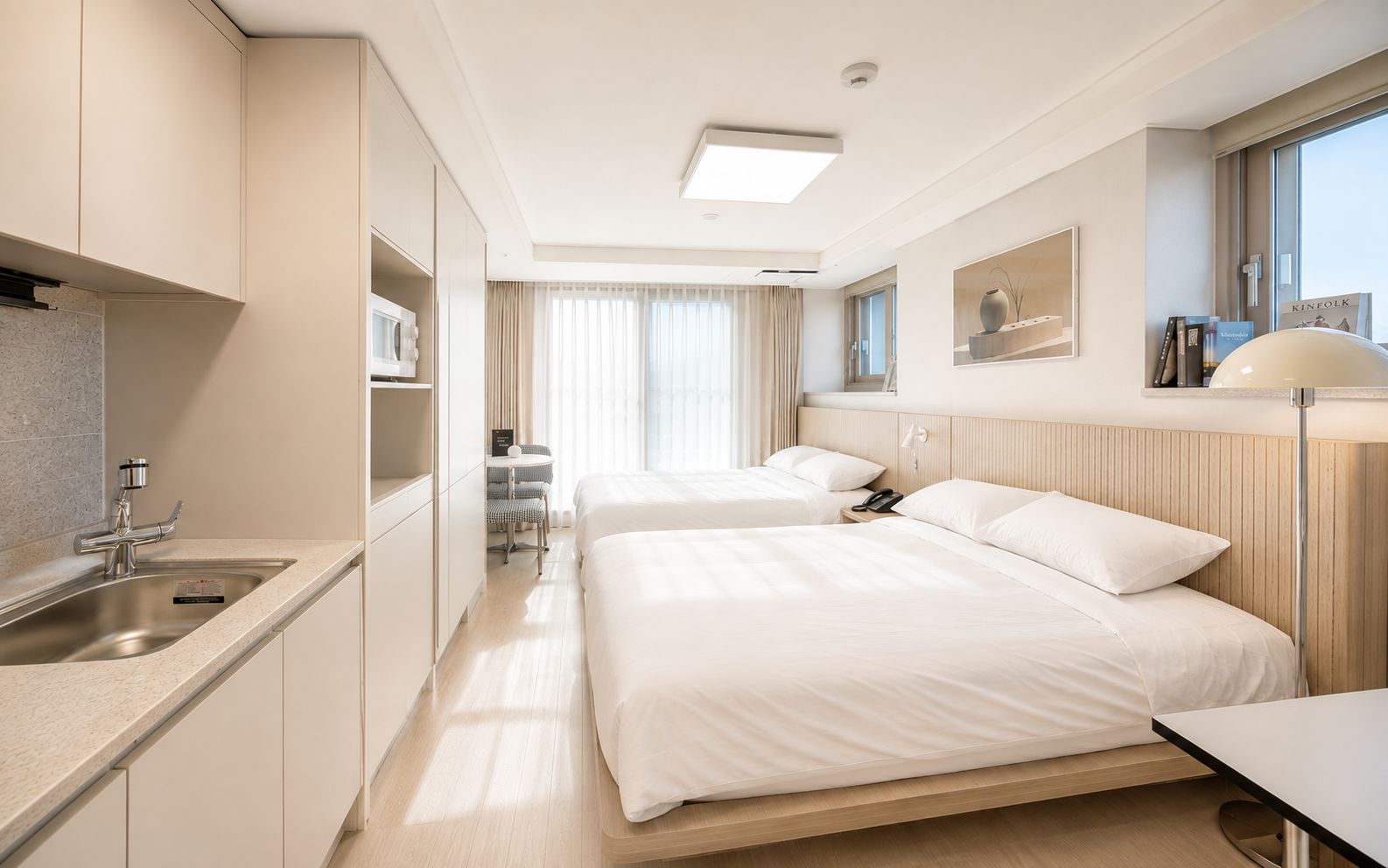 Ein Hotelzimmer mit Küchenzeile, zwei Betten, einem Nachttisch, einer Lampe und einem Fenster mit Vorhängen.
